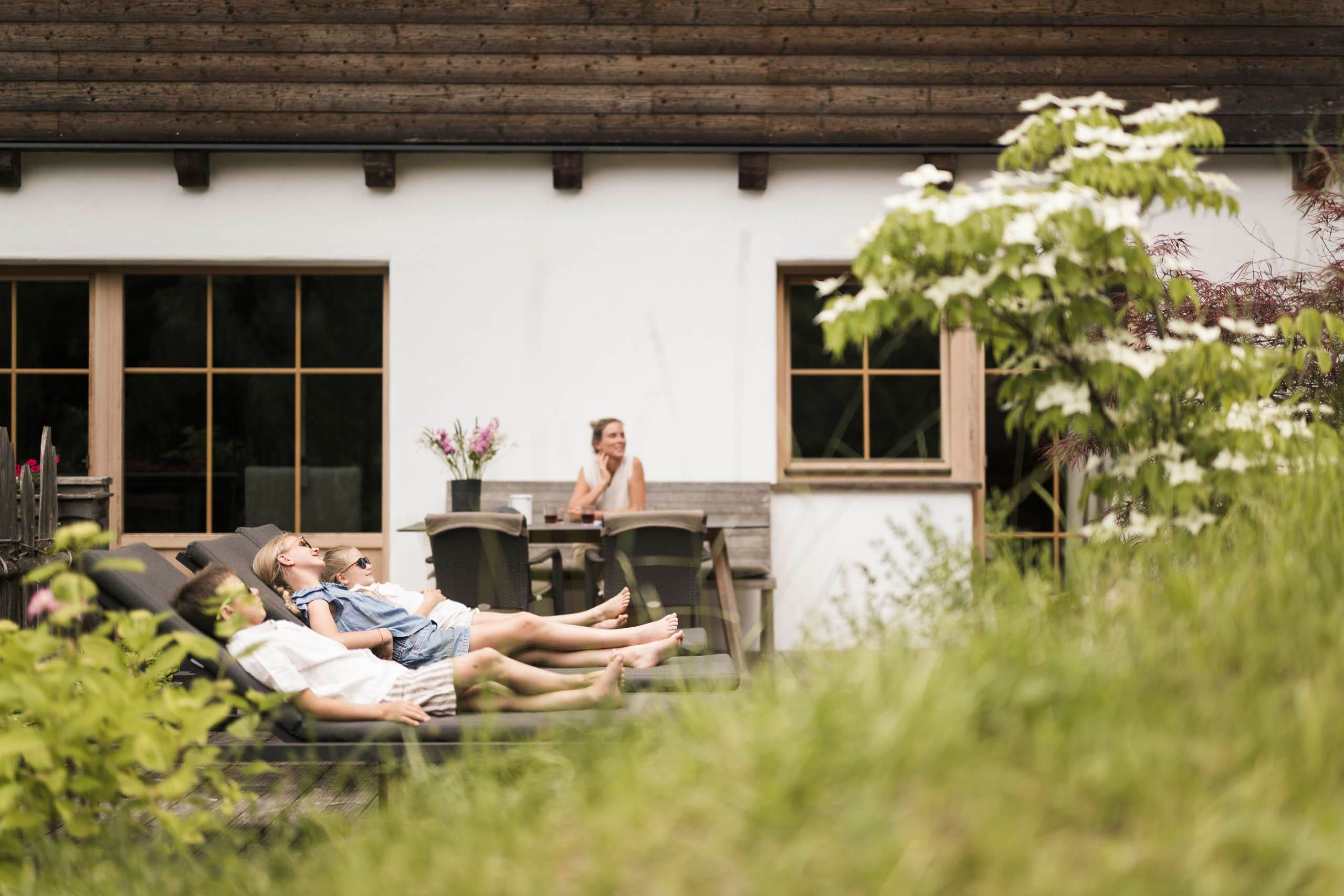 A family relaxes on lounge chairs in the garden of a house. In the background, a woman can be seen sitting in a chair and enjoying the surroundings.