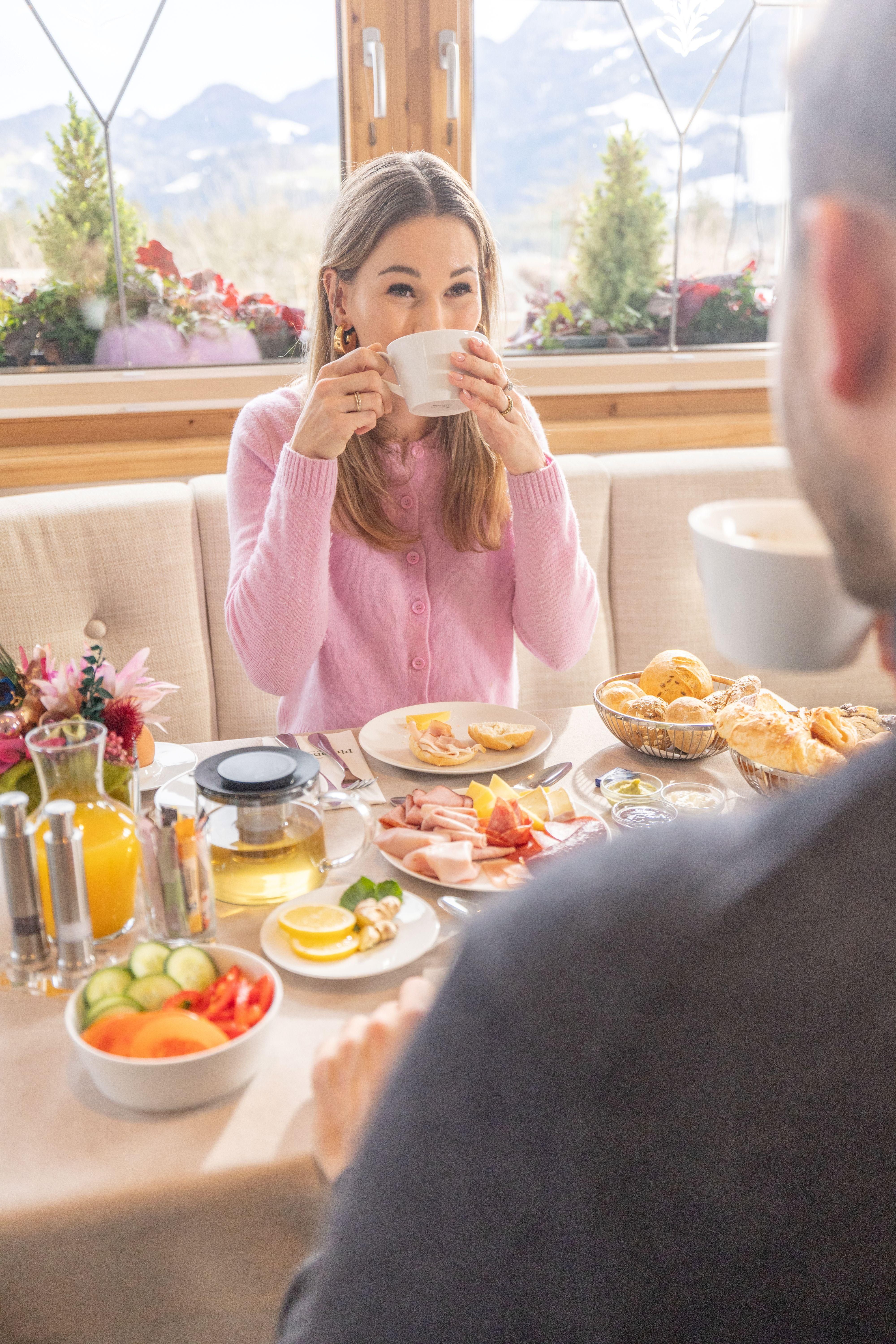 A breakfast table with various delicacies, including fruit, cold cuts, and rolls. A woman in a pink sweater is drinking a cup of tea or coffee and enjoying the beautiful view.
