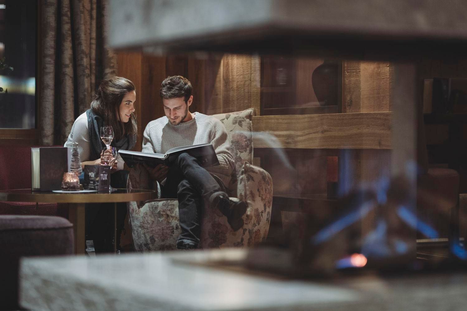 A couple sits comfortably in a modern room, looking at a menu together. In the background, a beautiful fireplace is burning, and a relaxed atmosphere can be felt.