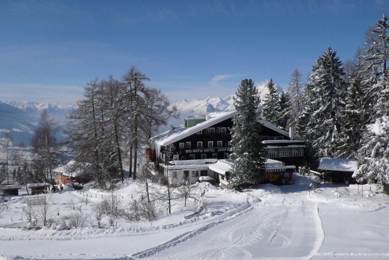A picturesque winter landscape with a chalet-like building, surrounded by snow-covered trees. In the background, majestic mountains and a clear sky can be seen.