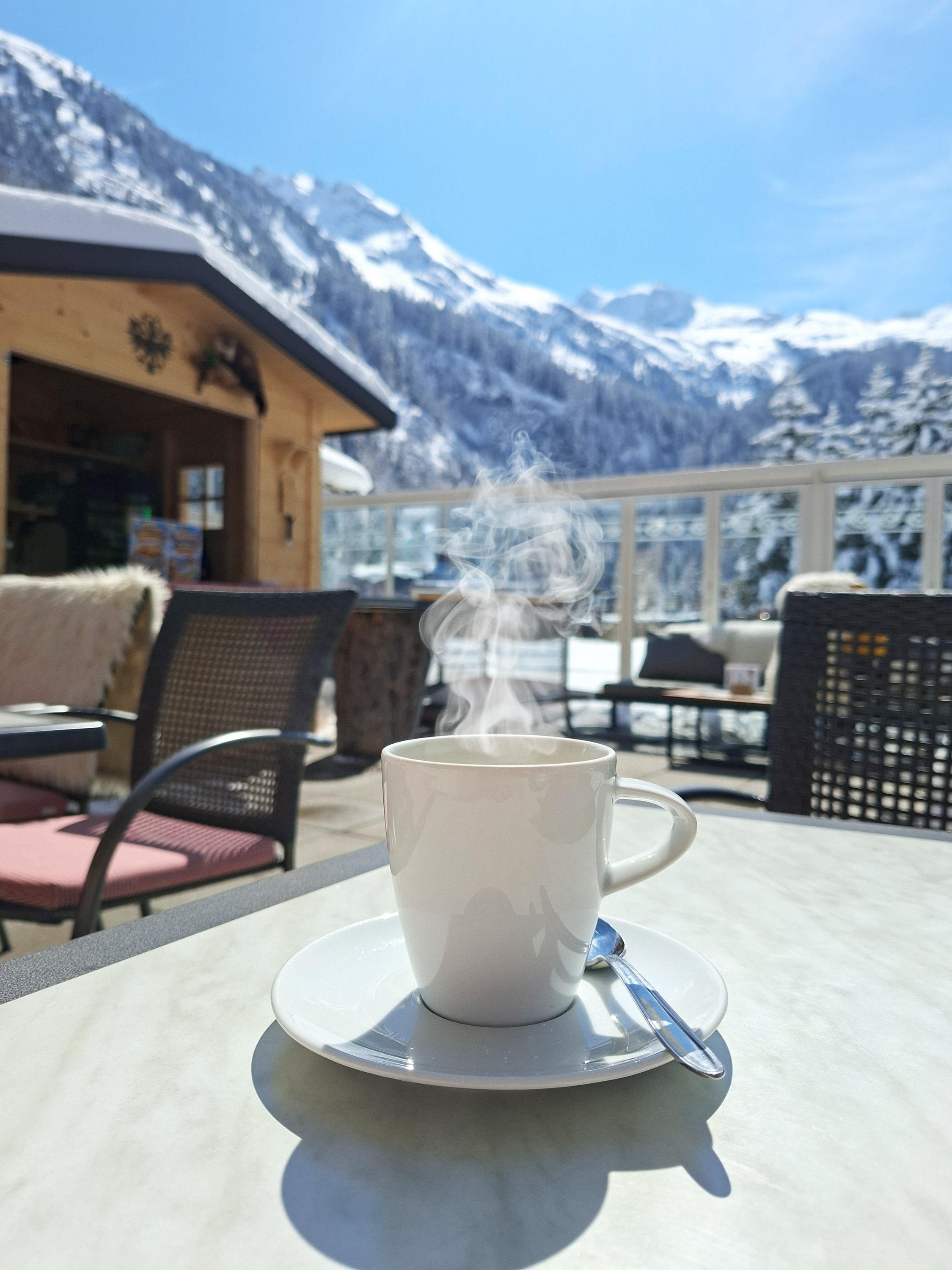 A steaming cup of coffee sits on a table. In the background, snow-covered mountains and a clear blue sky are visible.
