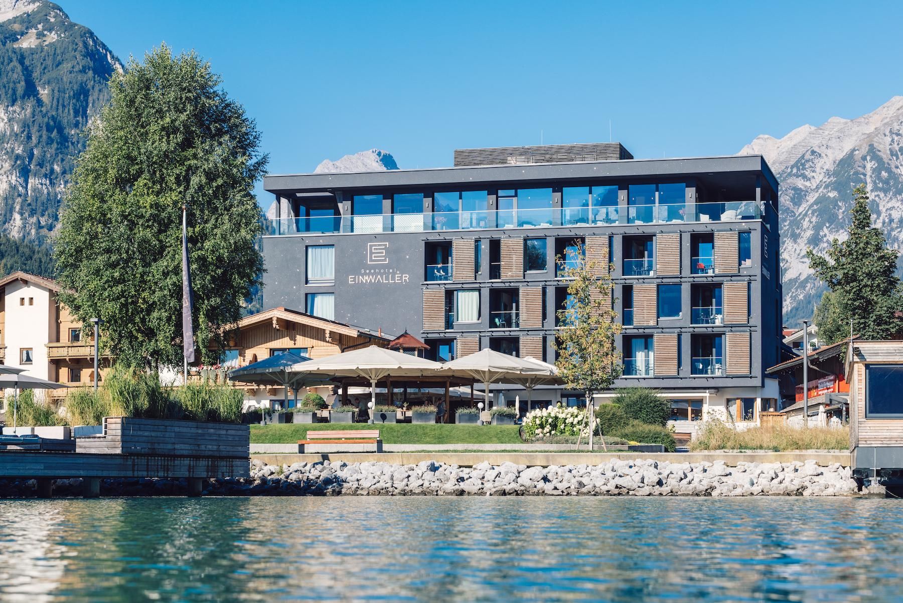 A modern hotel by the water with large windows and sun umbrellas. In the background, impressive mountains can be seen.