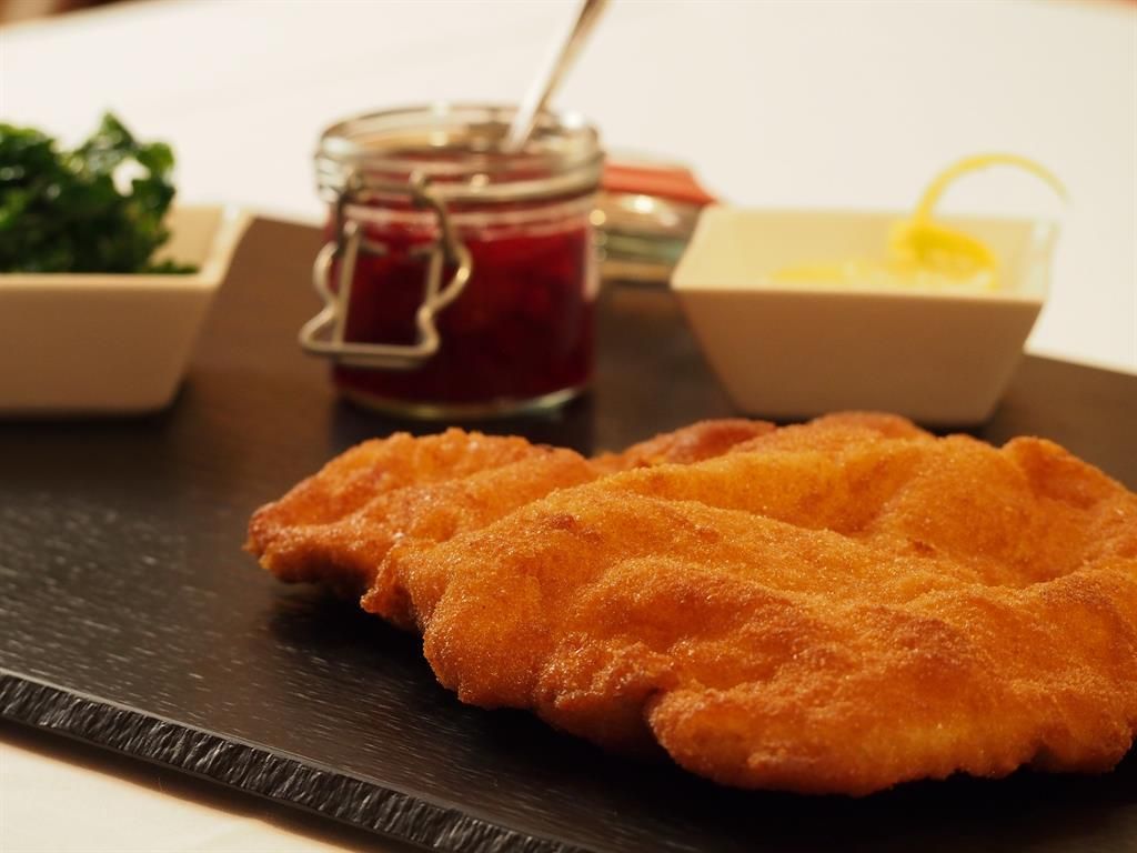 A crispy schnitzel on a black table. Next to it are sides like green leaves, jam, and a sauce.