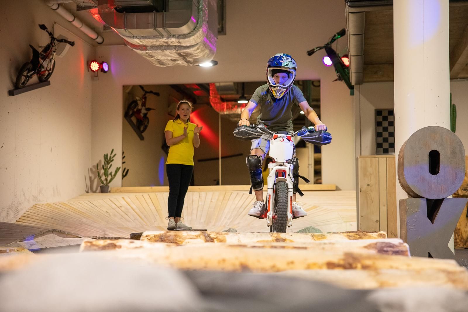 A boy on a motorcycle is navigating an obstacle course in an indoor sports facility. A woman in a yellow shirt is watching him critically.