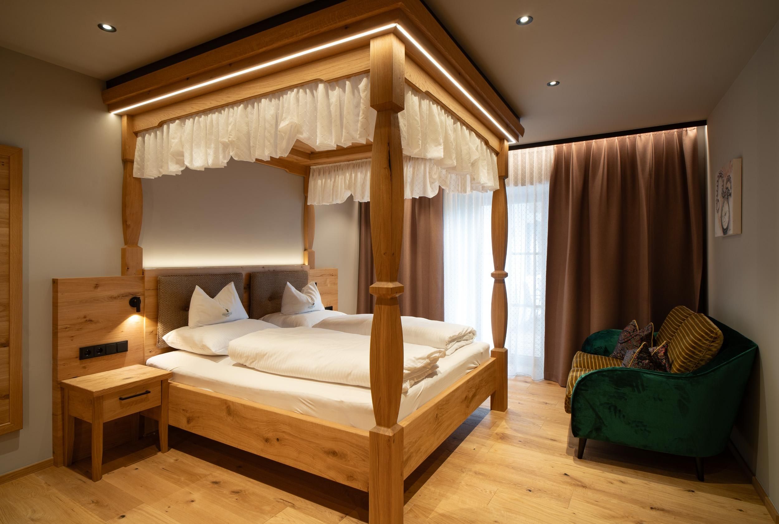 A cozy bedroom with a wooden four-poster bed. There is a small table and a green couch, as well as large windows with curtains.