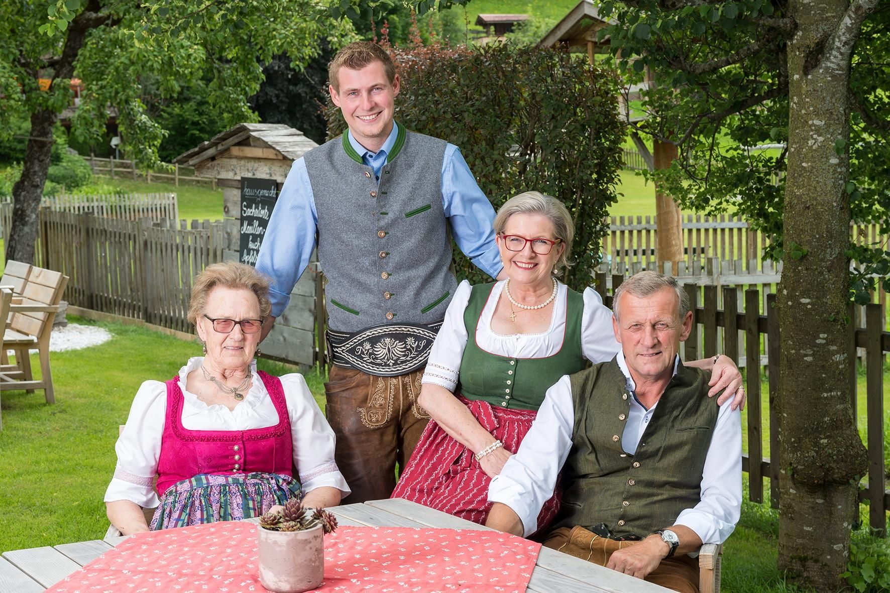 A group of four people in traditional attire is sitting at a table outdoors. In the background, there are trees and a well-maintained garden.