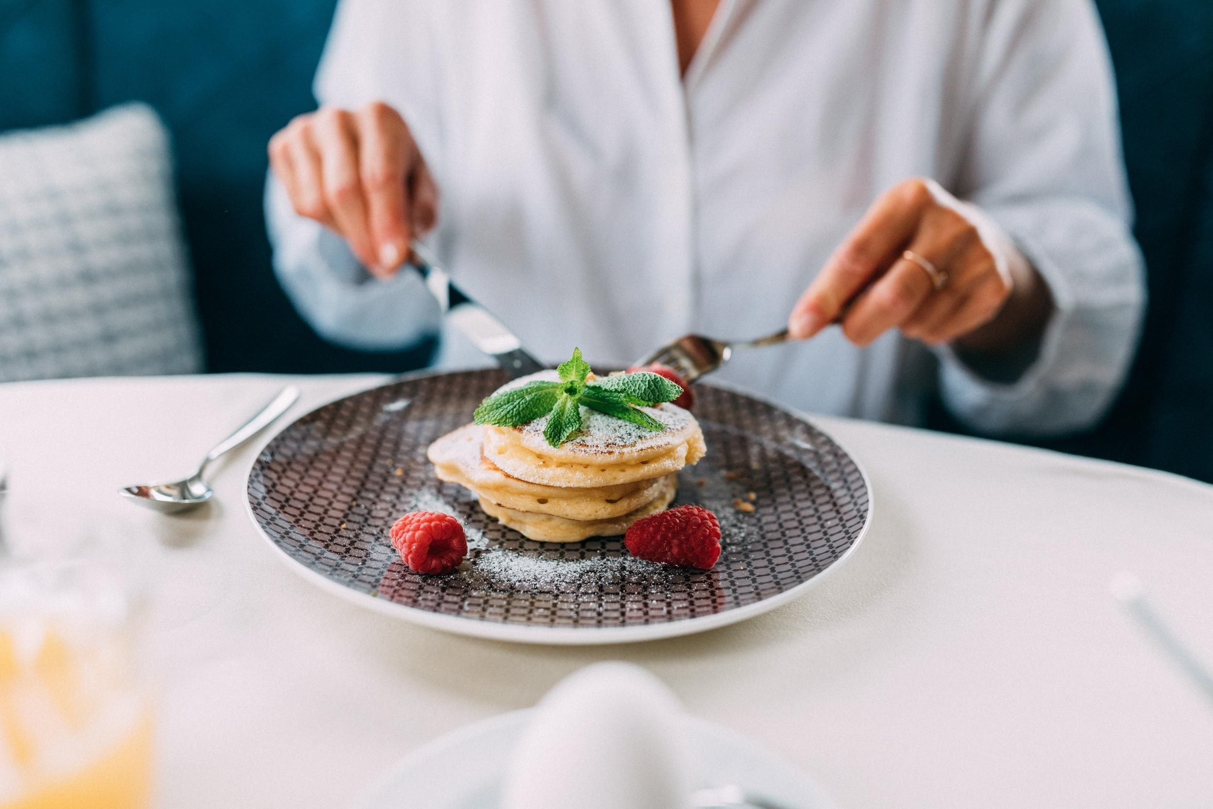 A plate with pancakes, garnished with fresh strawberries and mint. A person is cutting the pancakes with a knife and fork.
