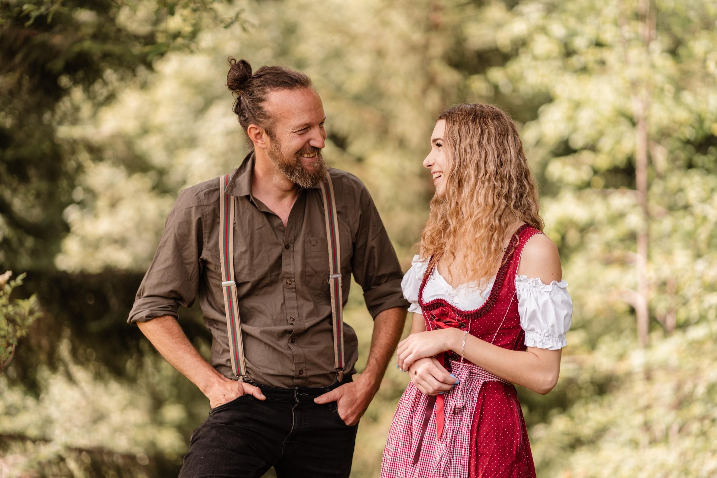 A smiling couple in traditional attire stands outdoors. Green trees can be seen in the background.