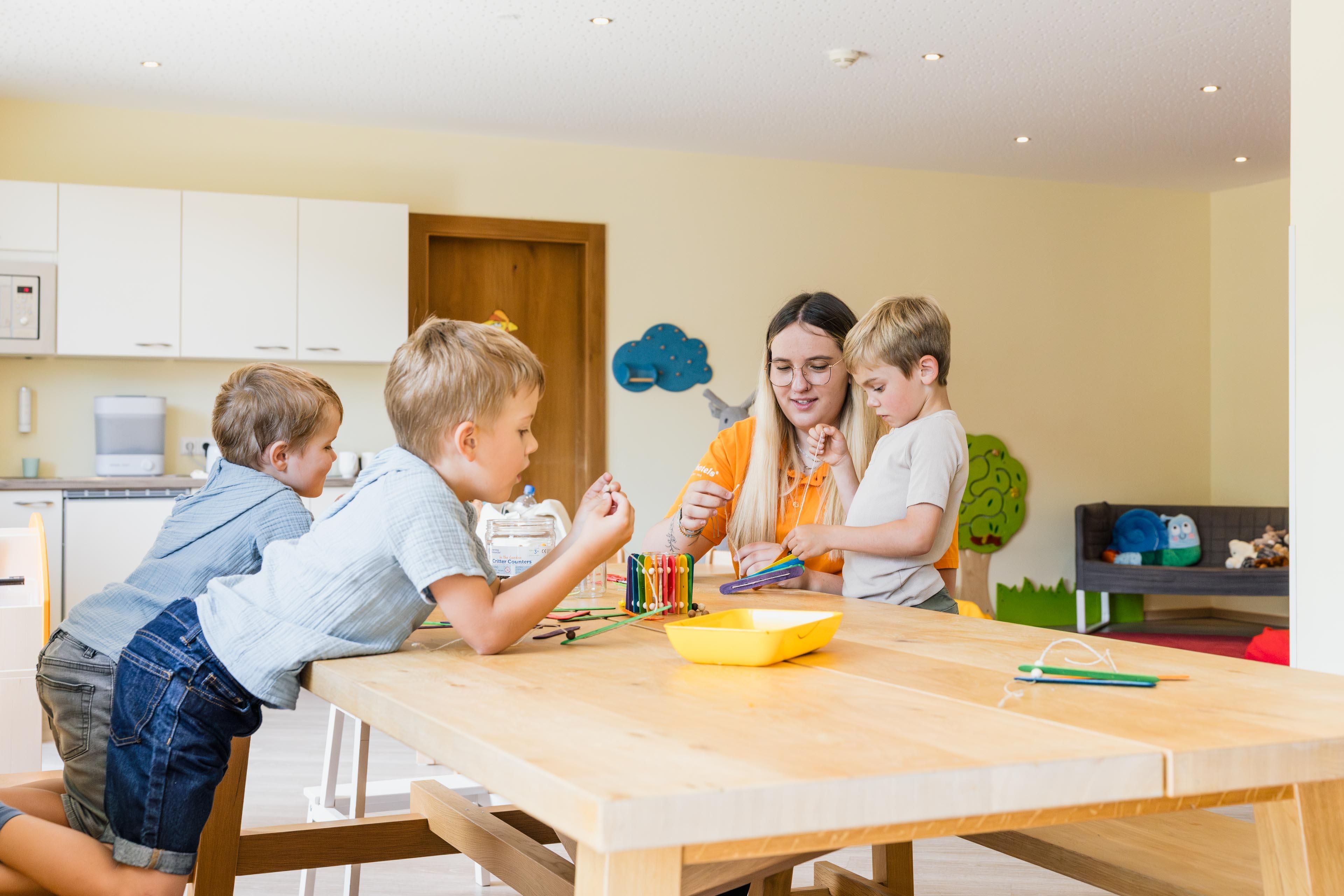 A room with a large table where children and a caregiver are crafting and being creative together. The environment is bright and friendly designed.