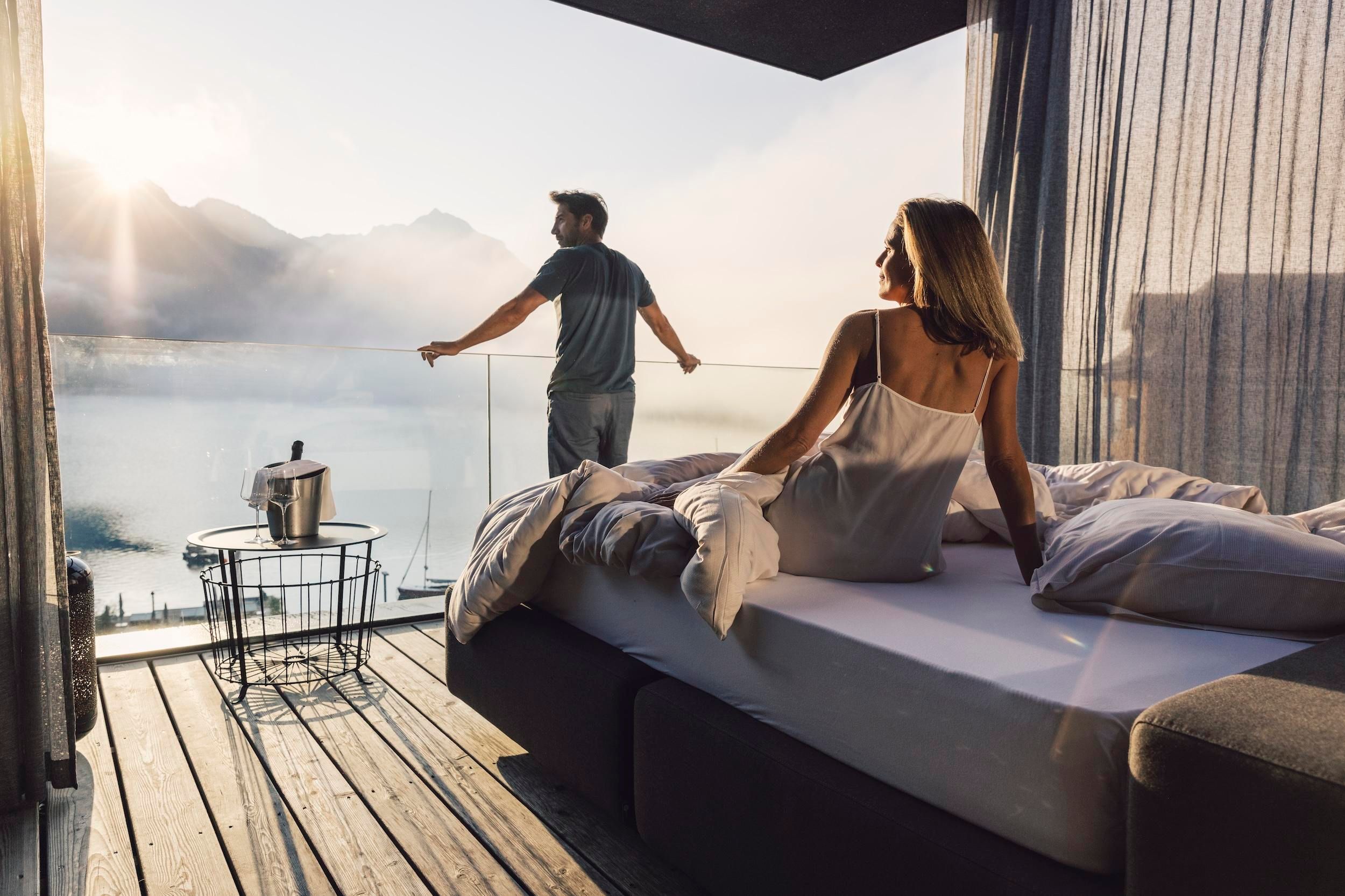 A couple enjoys the view of a picturesque lake from their modern bedroom. The atmosphere is tranquil and relaxing, with gentle rays of light shining through the windows.