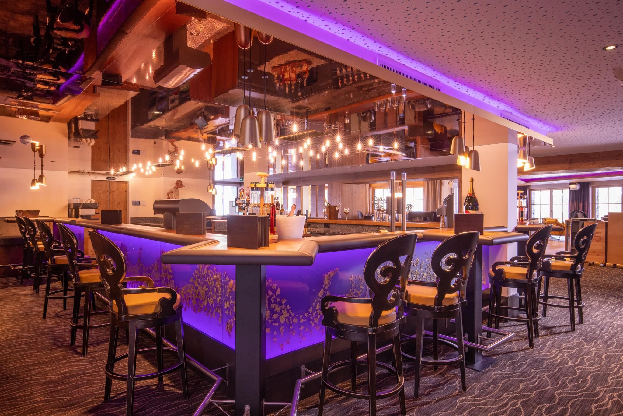 A modern bar with a brightly lit purple counter and elegant bar stools. The space is stylishly furnished and offers an inviting atmosphere.