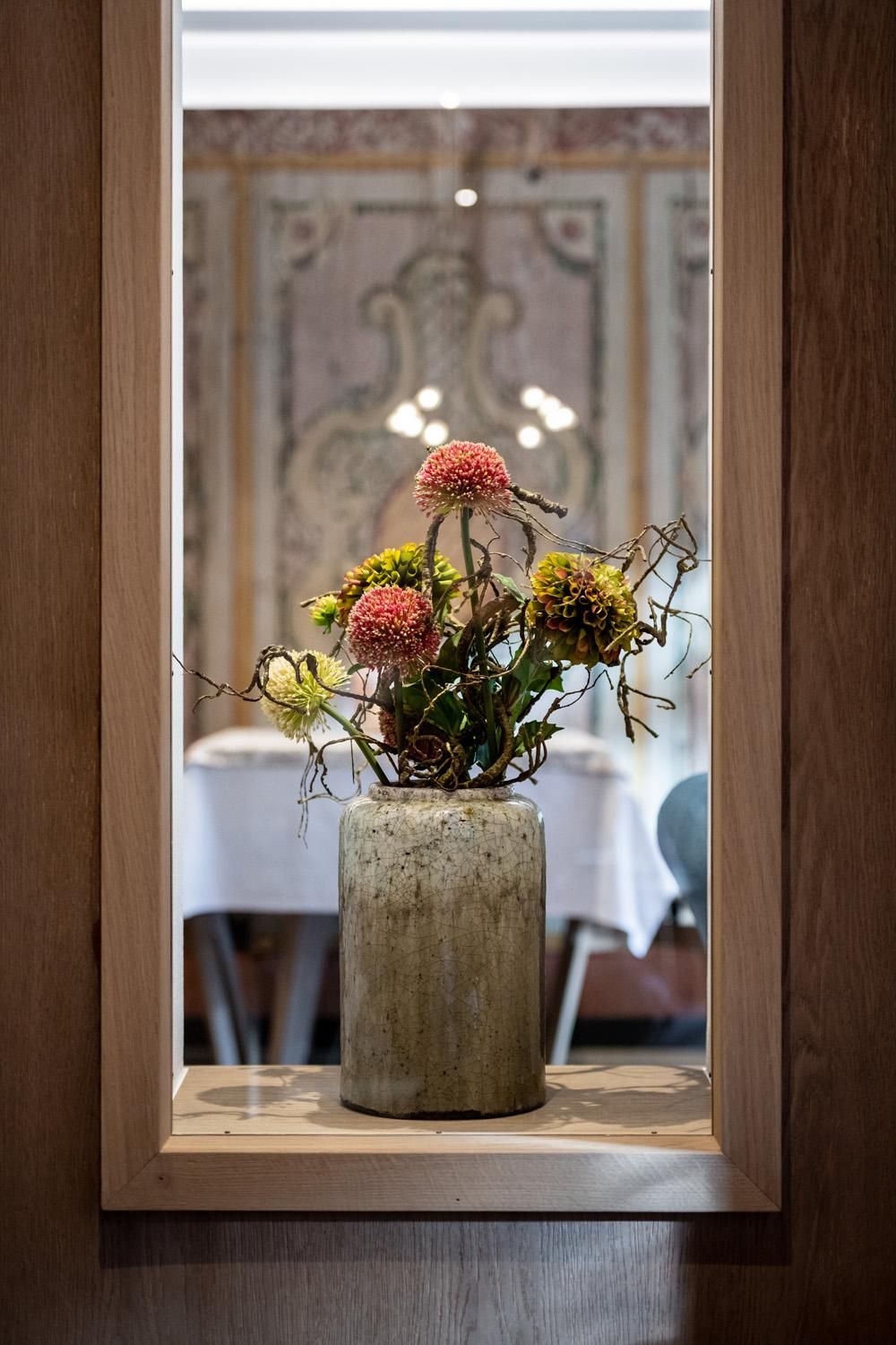 A flower arrangement in a gray vase stands behind a wooden frame. In the background, tables and an elegant wall design are visible.