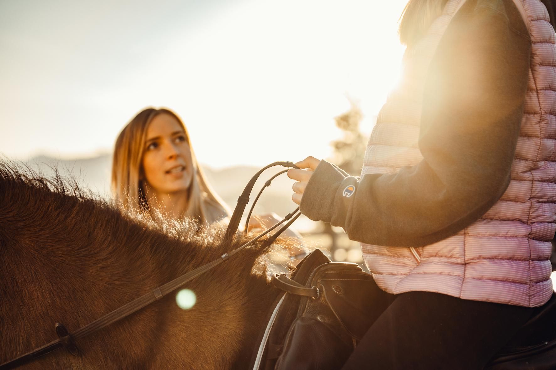 A woman is speaking with a rider on a horse. The sun is shining in the background, creating a warm atmosphere.