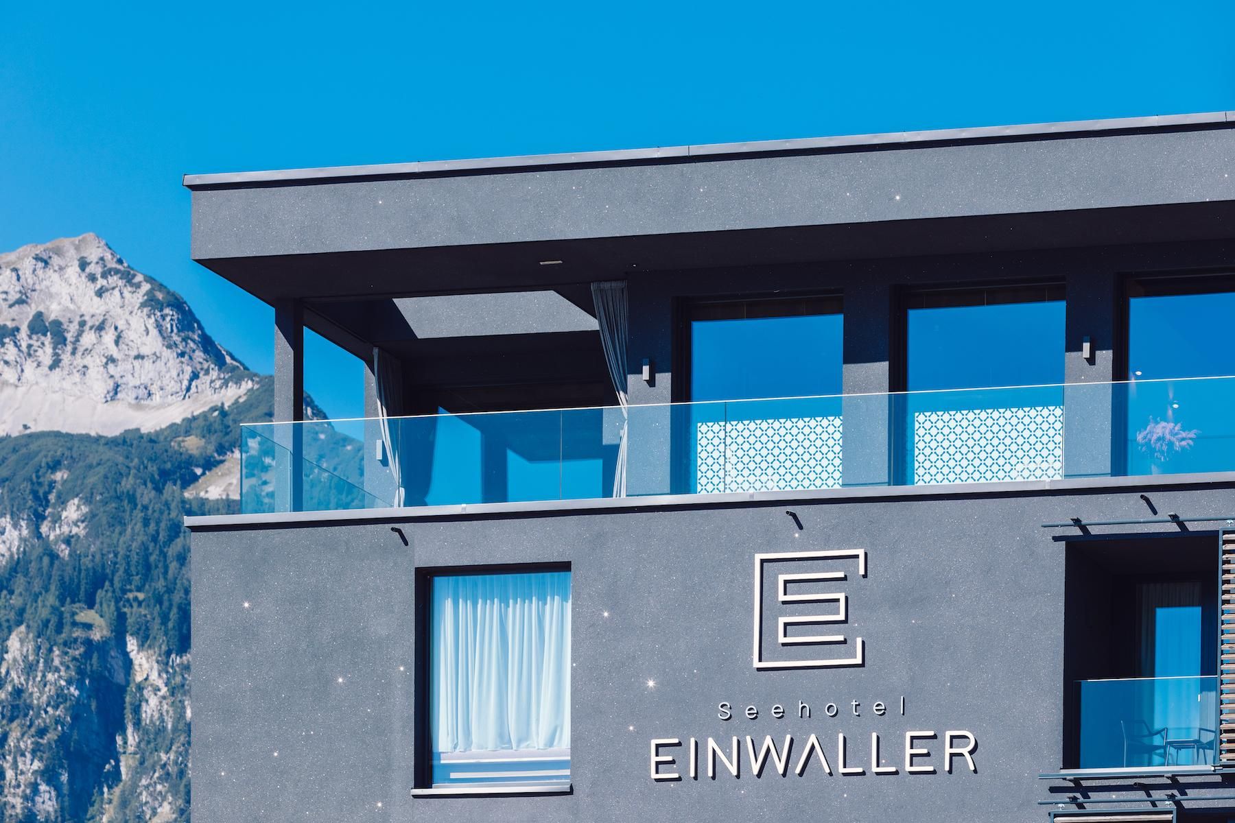 A modern hotel building named "EINWALLER". It is situated against a mountainous backdrop under a clear blue sky.