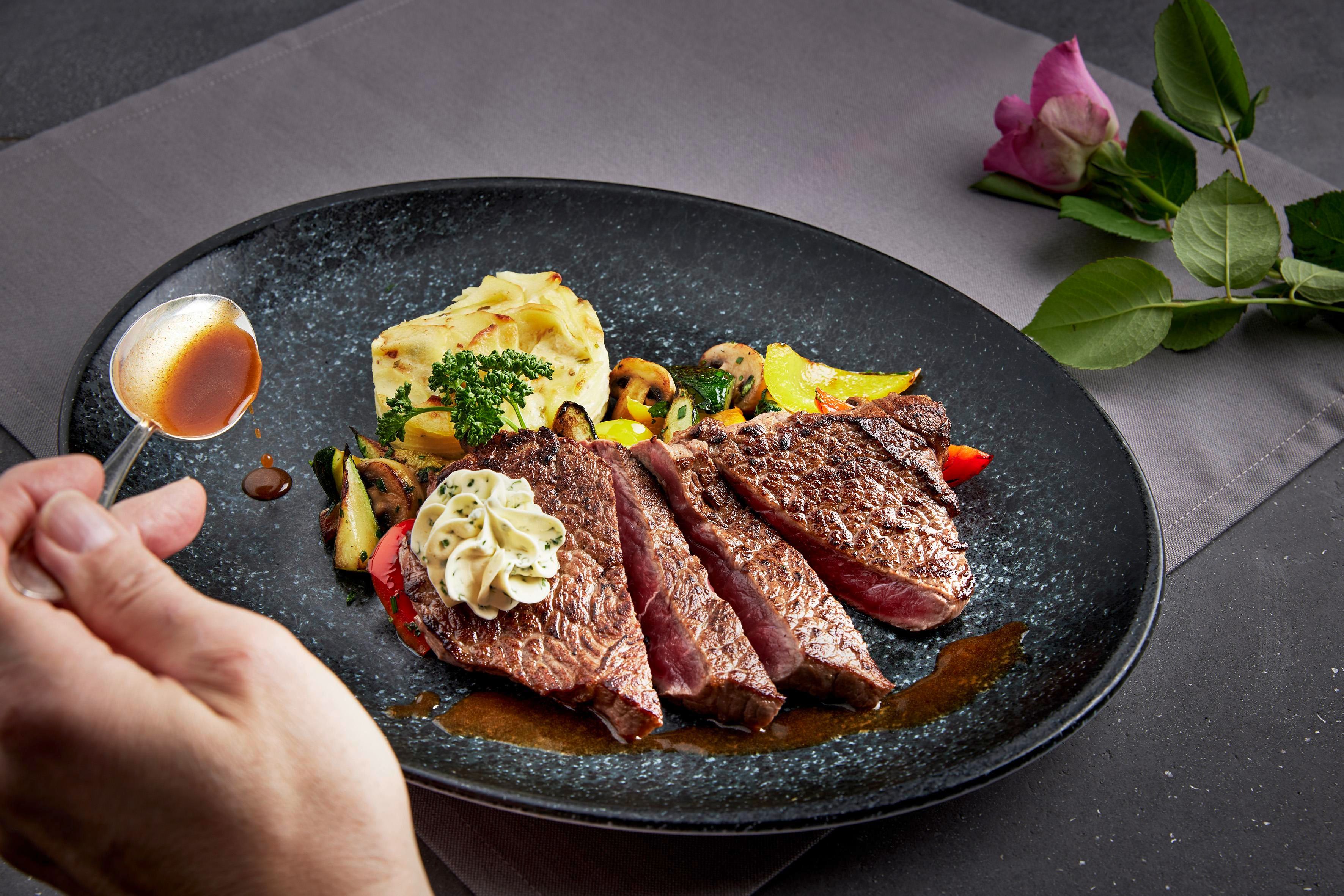 A plate with juicy meat, side dishes of vegetables, and a spoonful of sauce. An elegant presentation with a touch of flowers in the background.